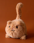 Soft stuffed animal in the shape of a round caramel colored cat with a question mark shaped tail
