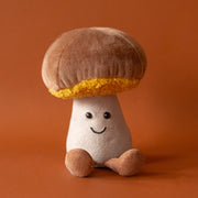 A soft suede stuffed animal in the shape of a mushroom with light brown cap with curly soft gold underneath, a tan colored stalk with smiley face and brown floppy legs.