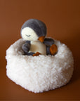 On a brown background is a penguin stuffed animal with a white boucle bed.