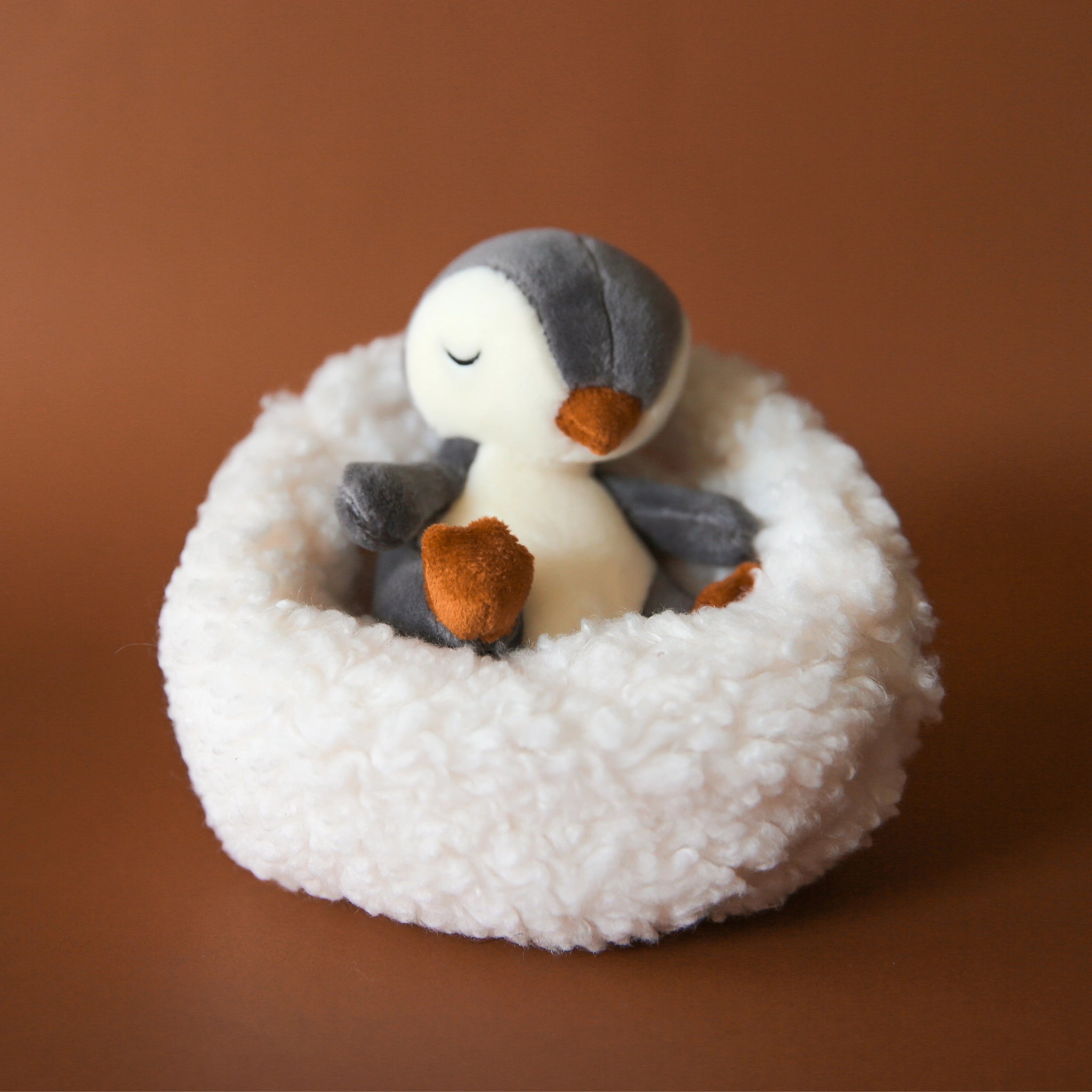 On a brown background is a penguin stuffed animal with a white boucle bed.