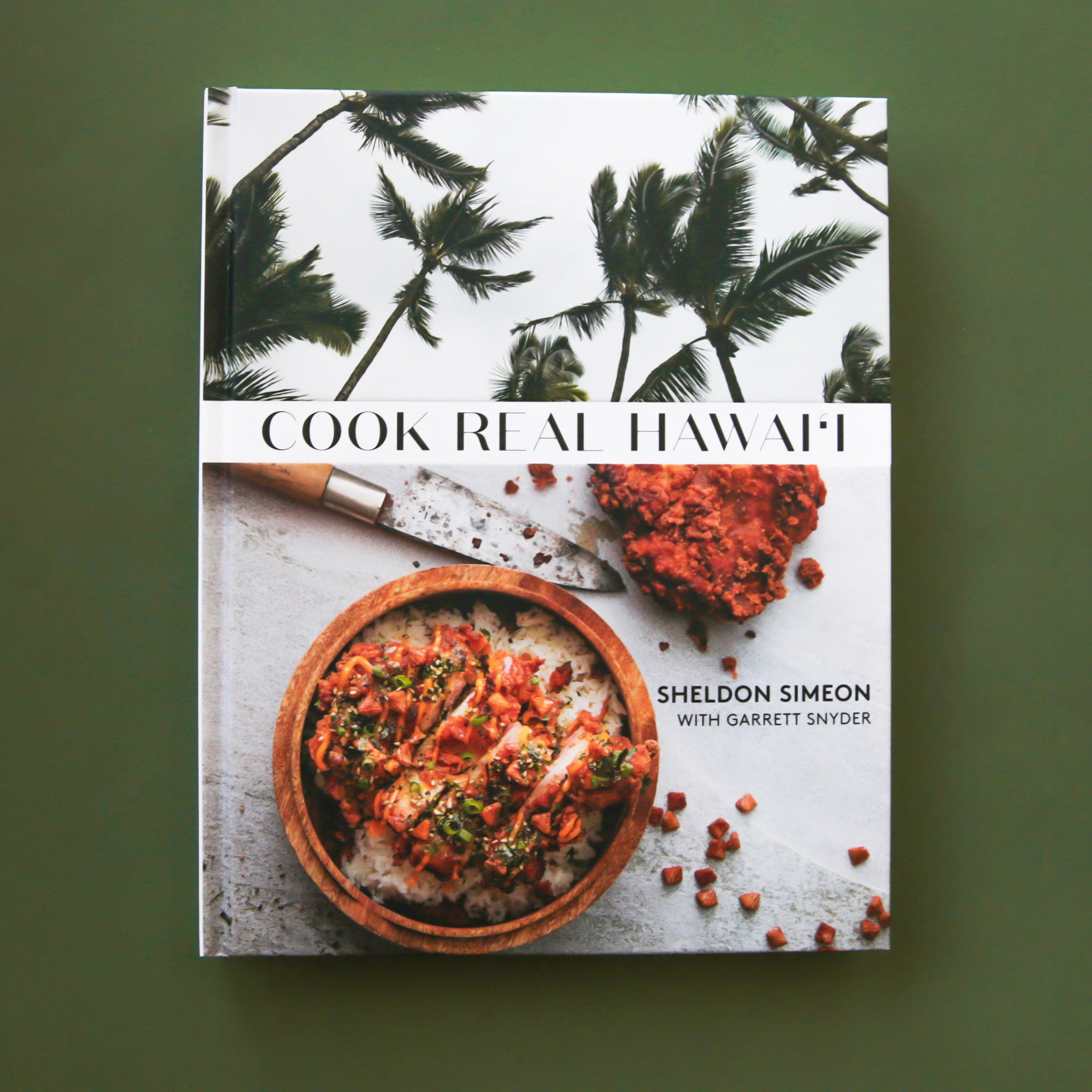 On a green background is a white book cover with palm trees and a vibrant meal in a bowl along with with text that reads, &quot;Cook Real Hawai&#39;i&quot;.