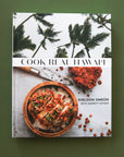 On a green background is a white book cover with palm trees and a vibrant meal in a bowl along with with text that reads, "Cook Real Hawai'i".