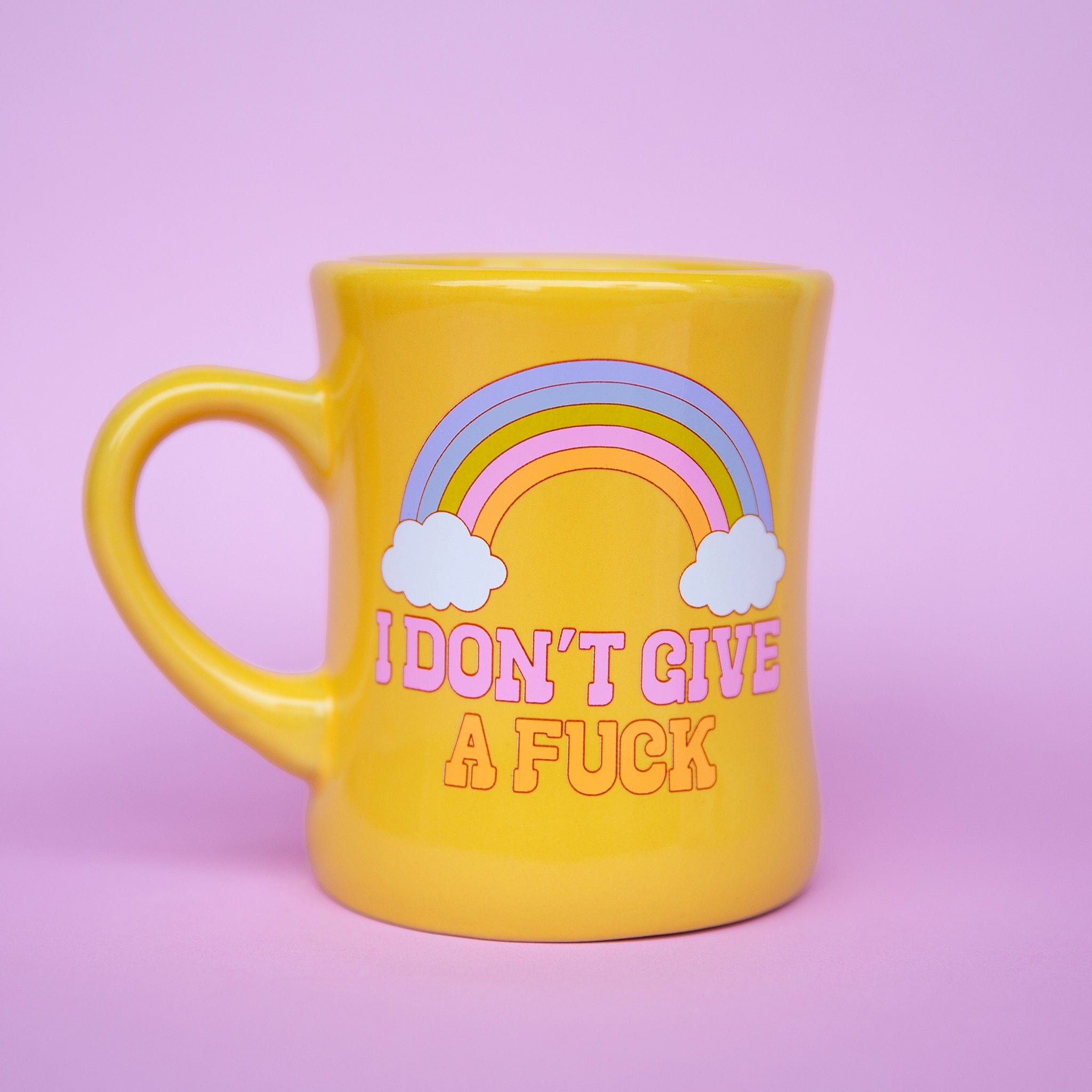 On a purple background is a yellow ceramic mug with a rainbow graphic as well as pink and orange text below it that reads, "I Don't Give A Fuck".