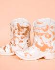 On a peachy background is two ceramic cowboy boot shaped candle with peach speckles on one and brown on the other.