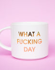 Classic white ceramic mug labeled 'What a Fucking Day' in gold shimmering capital lettering. Mug has a round white handle fasted on the left hand side.