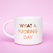 Classic white ceramic mug labeled 'What a Fucking Day' in gold shimmering capital lettering. Mug has a round white handle fasted on the left hand side.