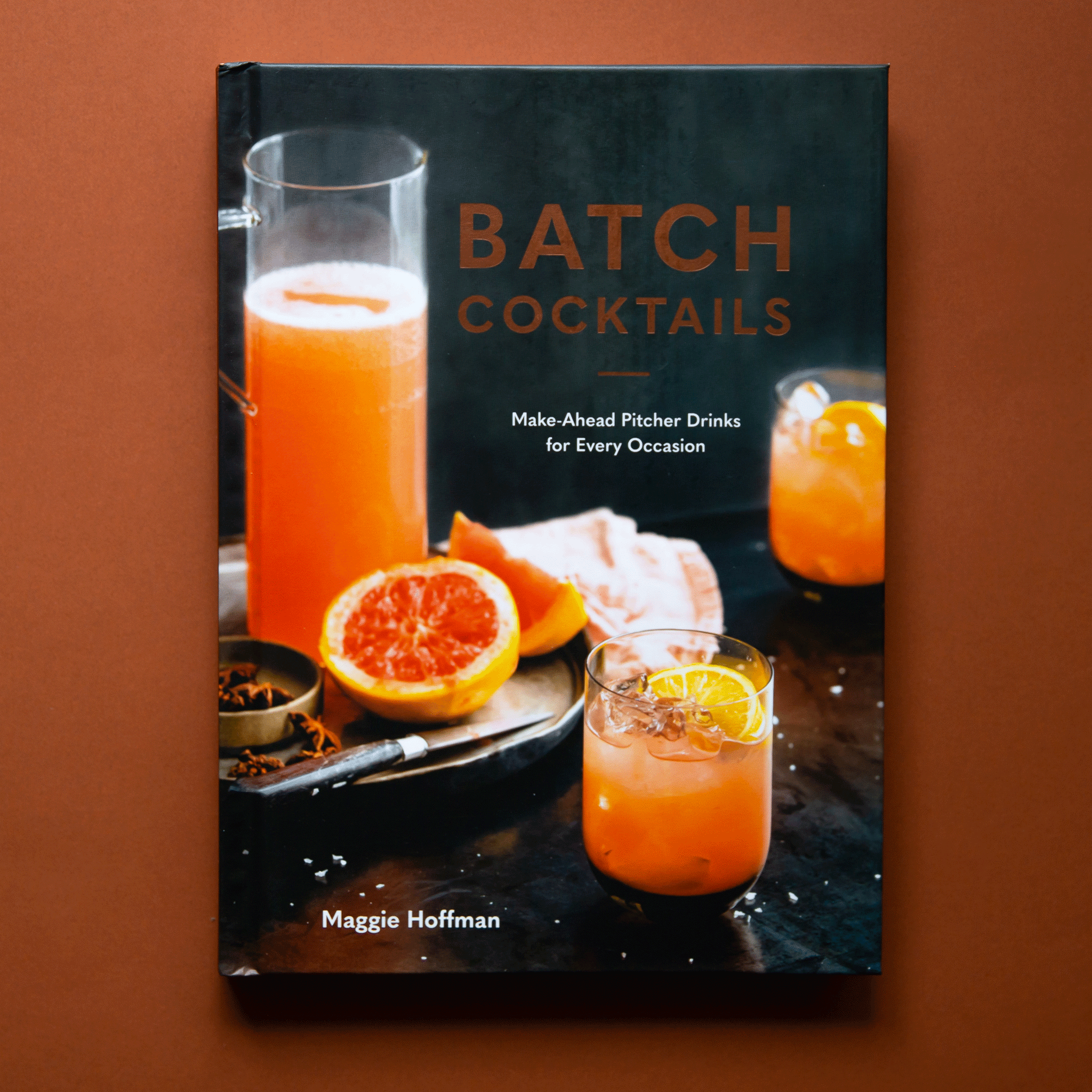 Cocktail book titled &quot;Batch Cocktails. Make-Ahead Pitcher Drinks for Every Occasion. Maggie Hoffman.&quot; The cover image shows a tray with a pitcher of tangerine liquid, a sliced blood orange, and a knife. Two tangerine colored cocktails with lemon garnishes sit beside the tray.