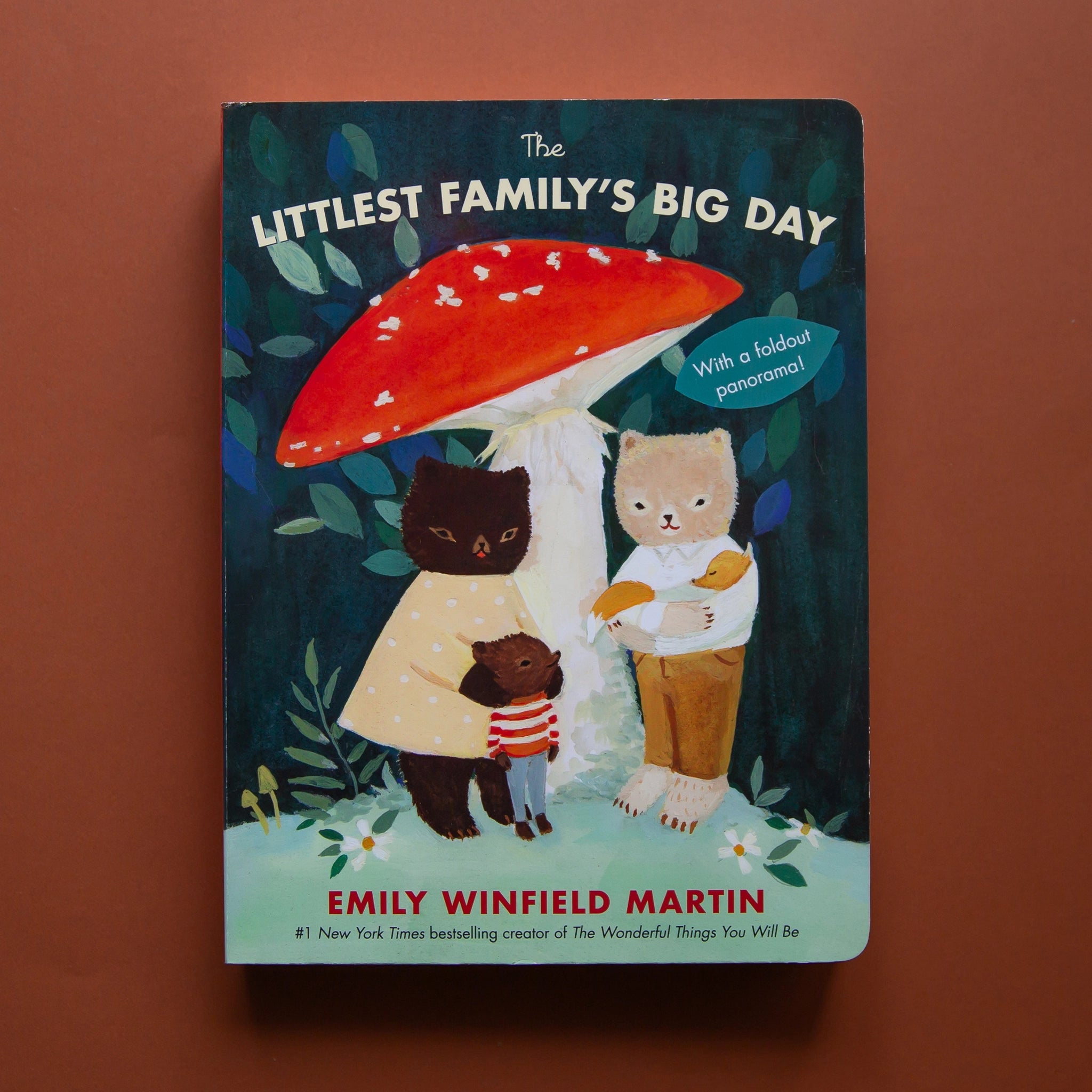 On a brown background is a green children's book cover with a small bear family standing under a red mushroom and the title above it that reads, "The Littlest Family's Biggest Day".