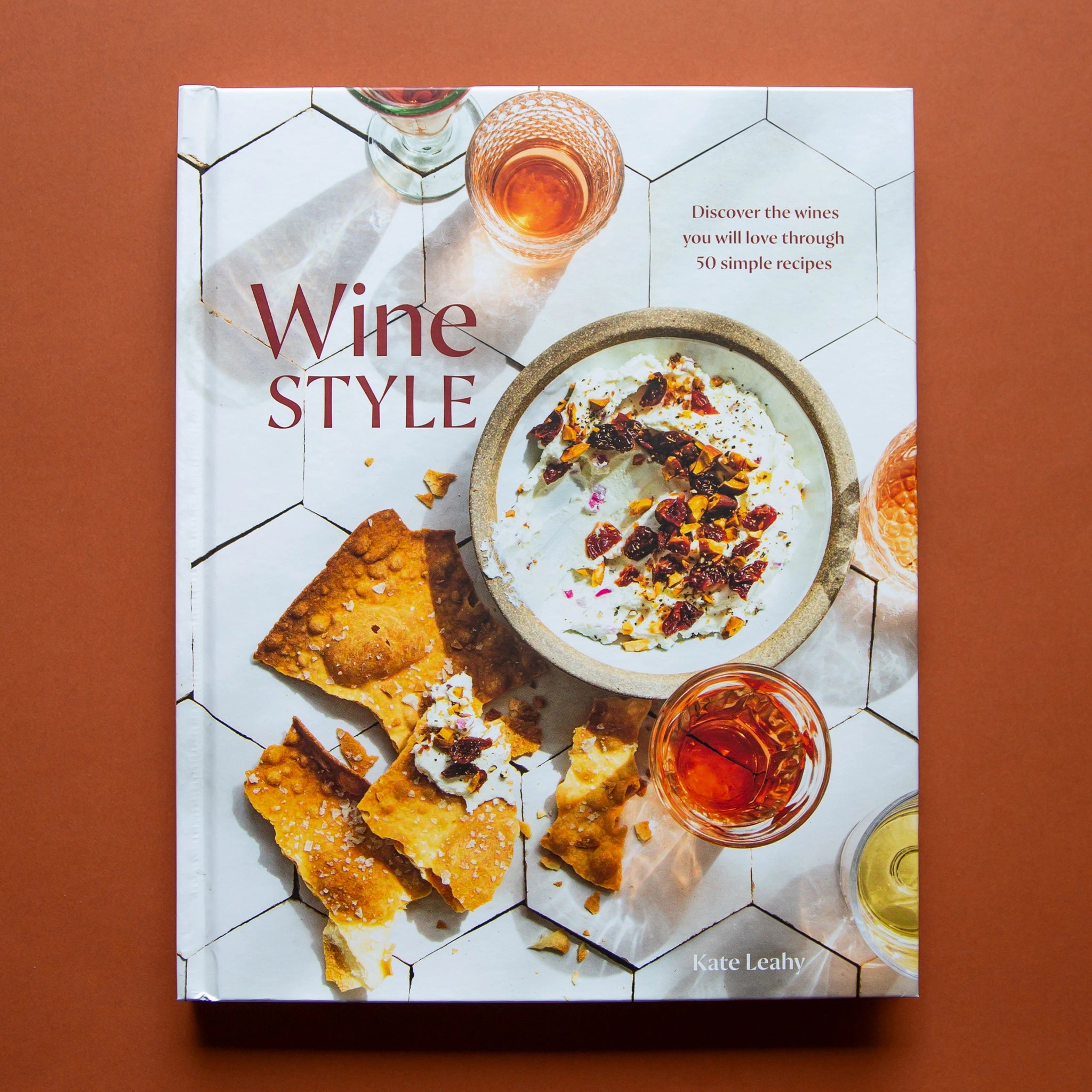 Hard covered cookbook titled &#39;Wine Style: Discover the wines you will love through 50 simple recipes&#39; in red lettering. Book cover features brittle salted crackers and dip along side wine-filled cocktail glasses against white modern tiled.