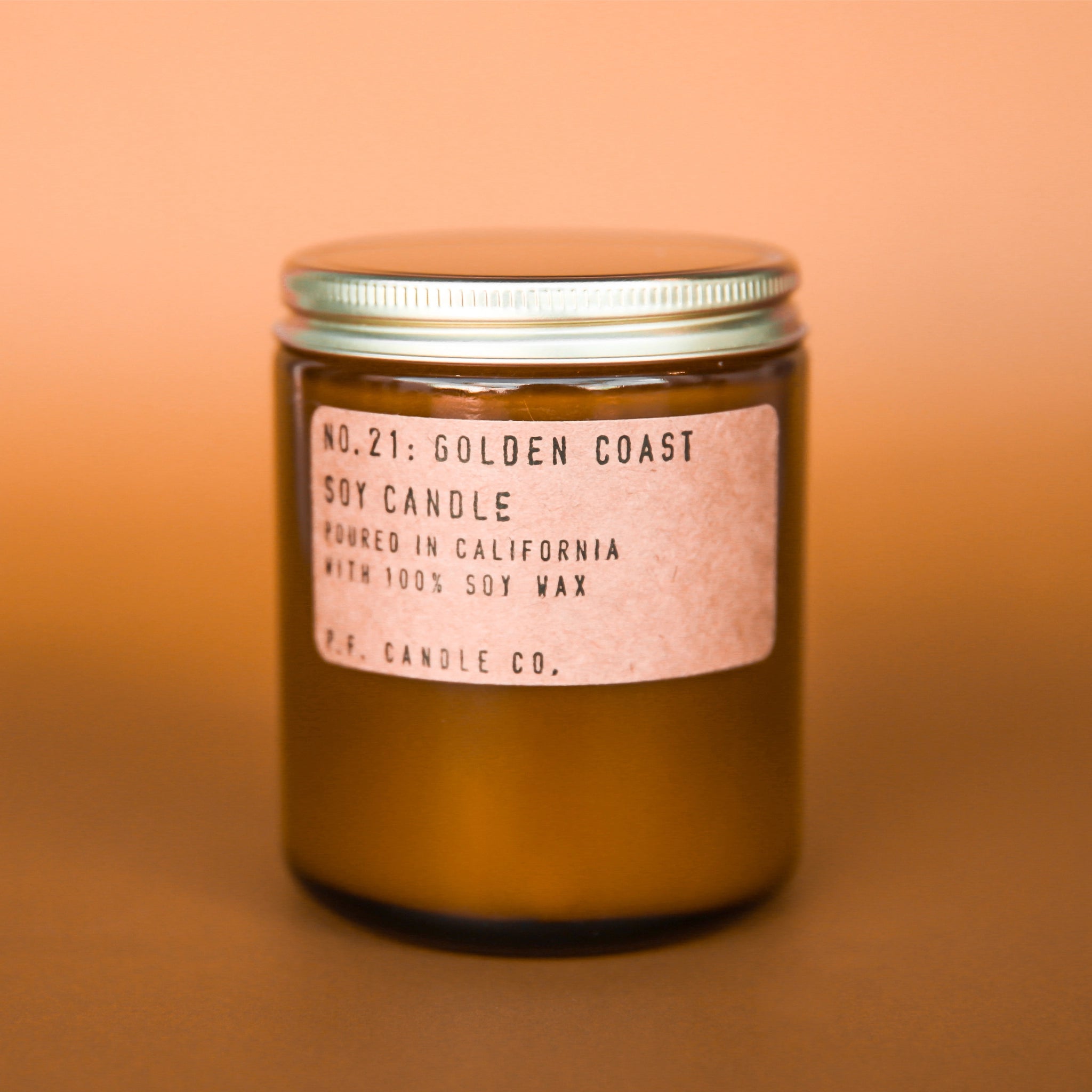 Candle features amber glass with a gold lid. The label is kraft paper with typewriter font that reads &quot;No.21: Golden Coast Soy Candle Handmade in California with 100% Soy Wax. P.F. Candle Co.&quot;