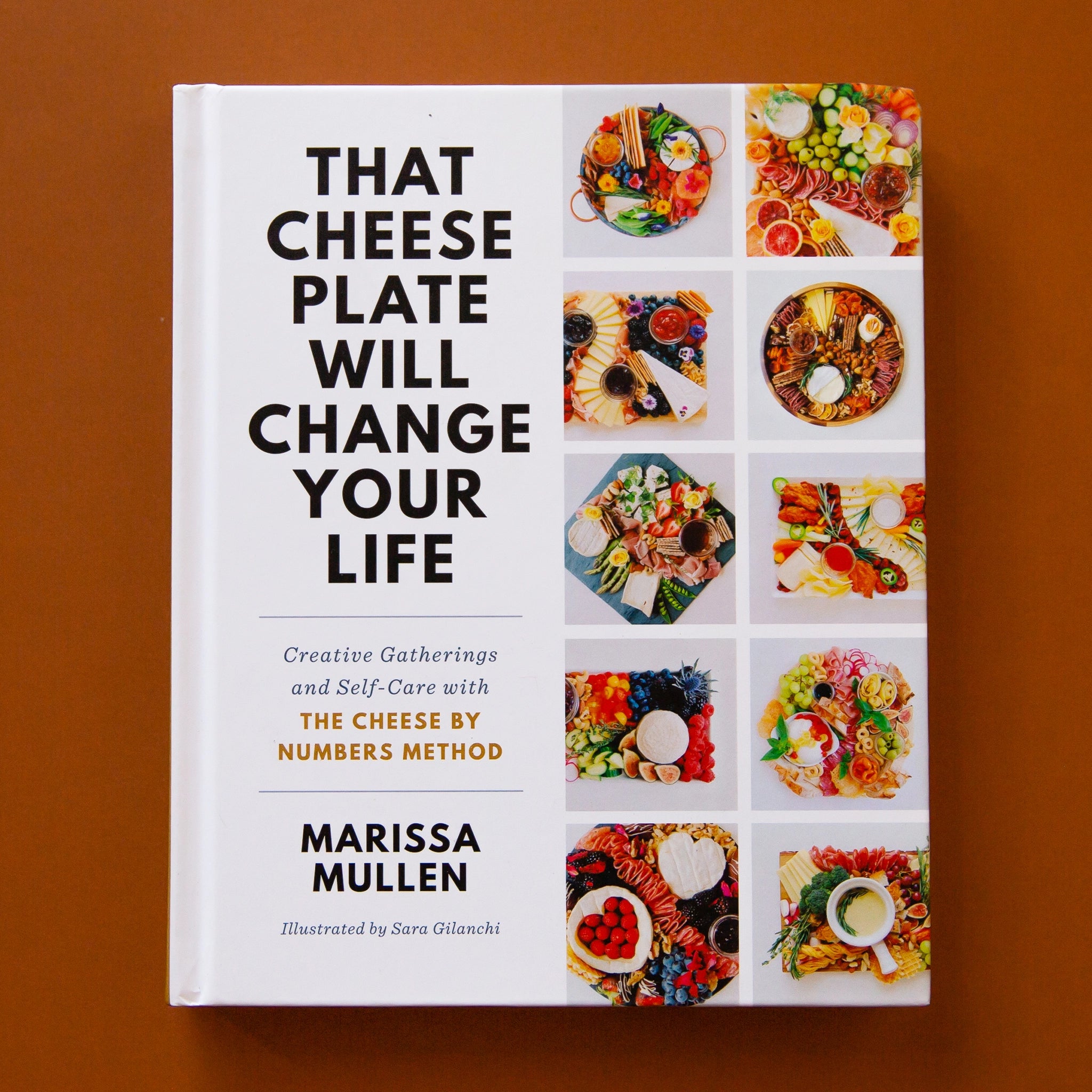 The front cover of the book reads &#39;That Cheese Plate Will Change Your Life&#39; in bold black lettering on the left side. Below reads &#39;Creative Gathering and Self-Care with The Cheese by Numbers Method&#39; in smaller text. To the right is 10 squares filled with a variety of well put together charcuterie boards, filled with cheeses, crackers, jams and more.