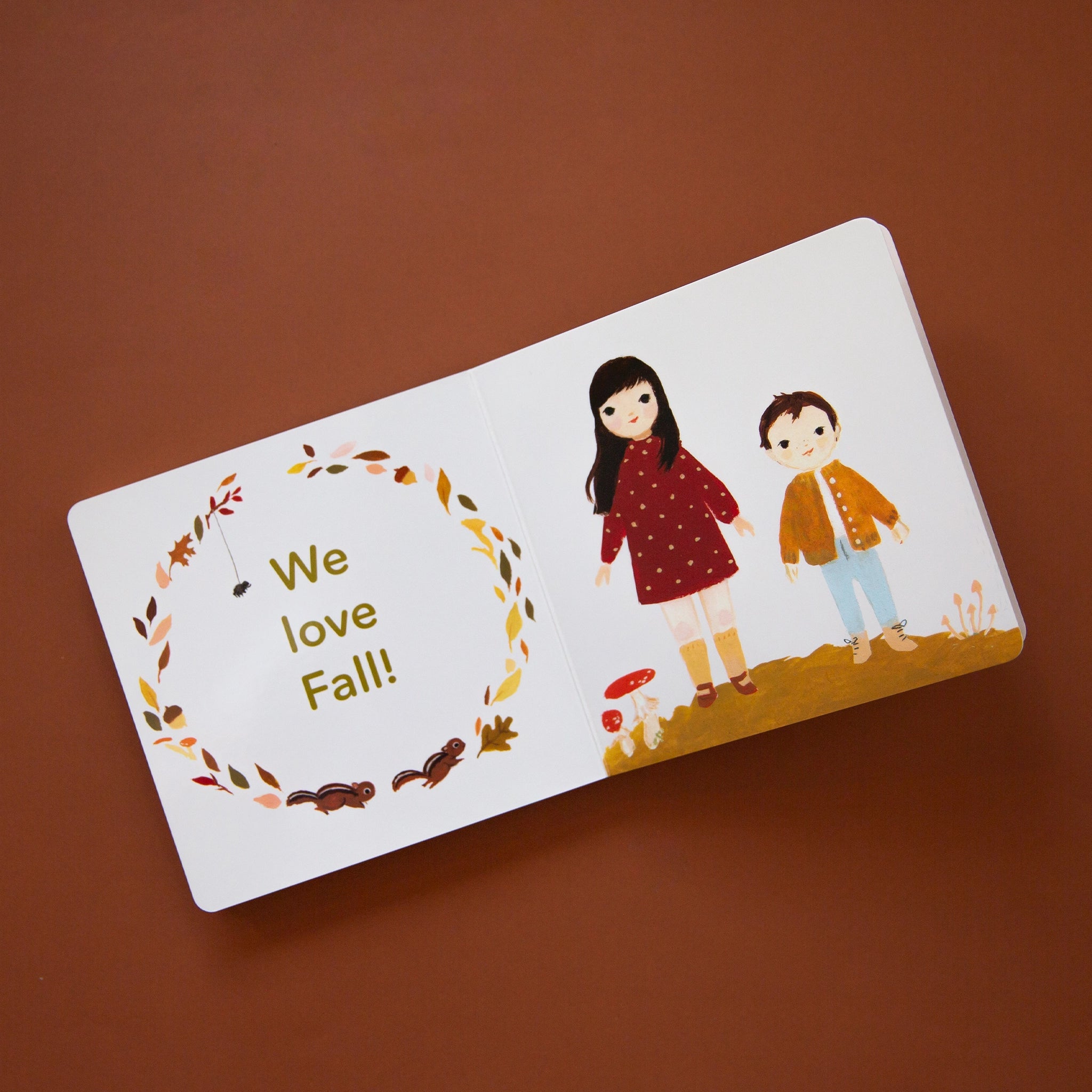 On a brown background is children's picture book is open to a page that has fall leaves and chipmunks in a circle around text that reads, "We love Fall!" as well as two children on the right page wearing warm toned fall sweaters and dresses. 