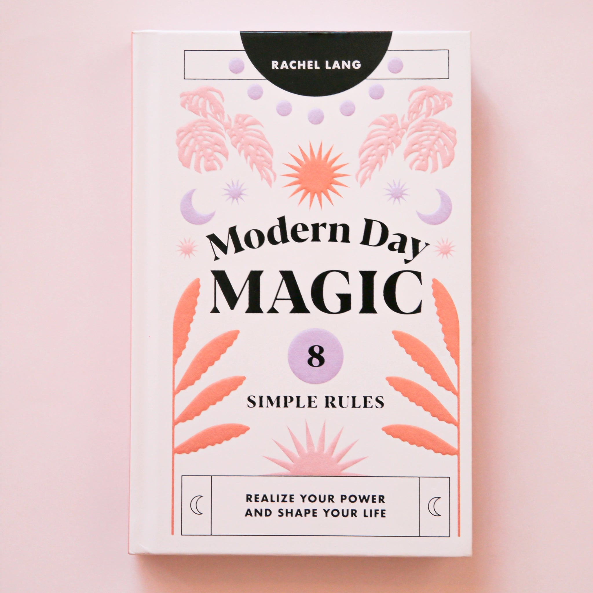 A light pink book cover with pink, lavender and orange foliage and sun and moon graphics along with black text that reads, &quot;Modern Day Magic 8 Simple Rules Realize Your Power and Shape Your Life&quot;.