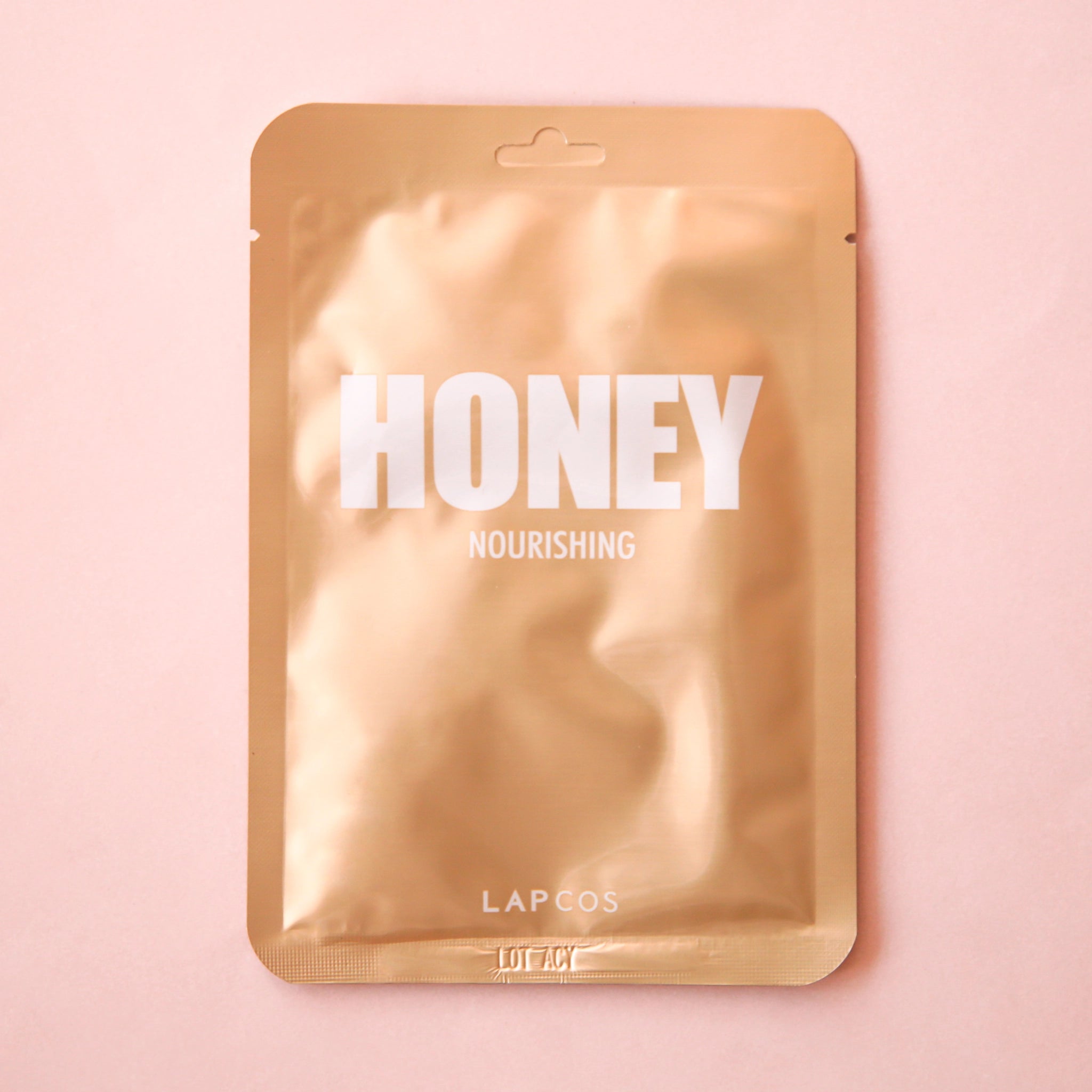 On a pink background is a gold packet holding a face mask with white text across the front that reads, &quot;Honey Nourishing&quot;. 
