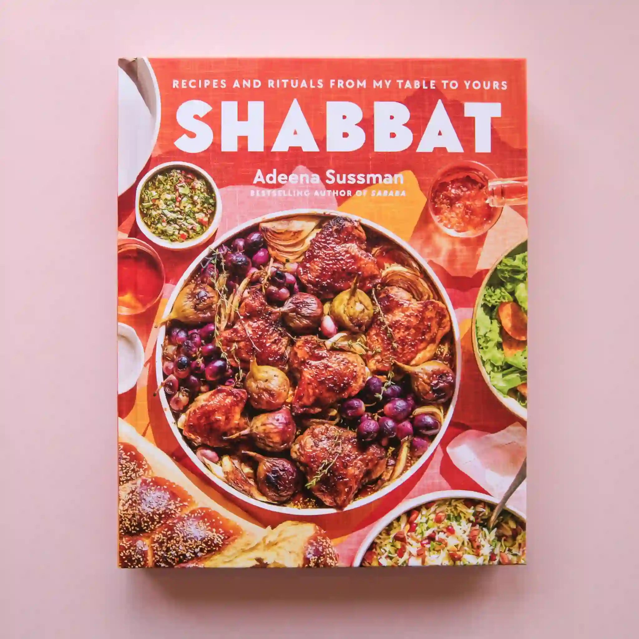 On a pink background is a colorful book cover with a big family style meal with white text at the top that reads, "Recipes and RItuals From My Table To Yours Shabbat".