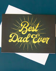 On a blue background is a dark greenish black Father's Day card with gold text across the front that reads, "Best Dad Ever".