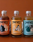 On a brown background is a modern cocktail kit with three glass bottles of cocktail mixes.