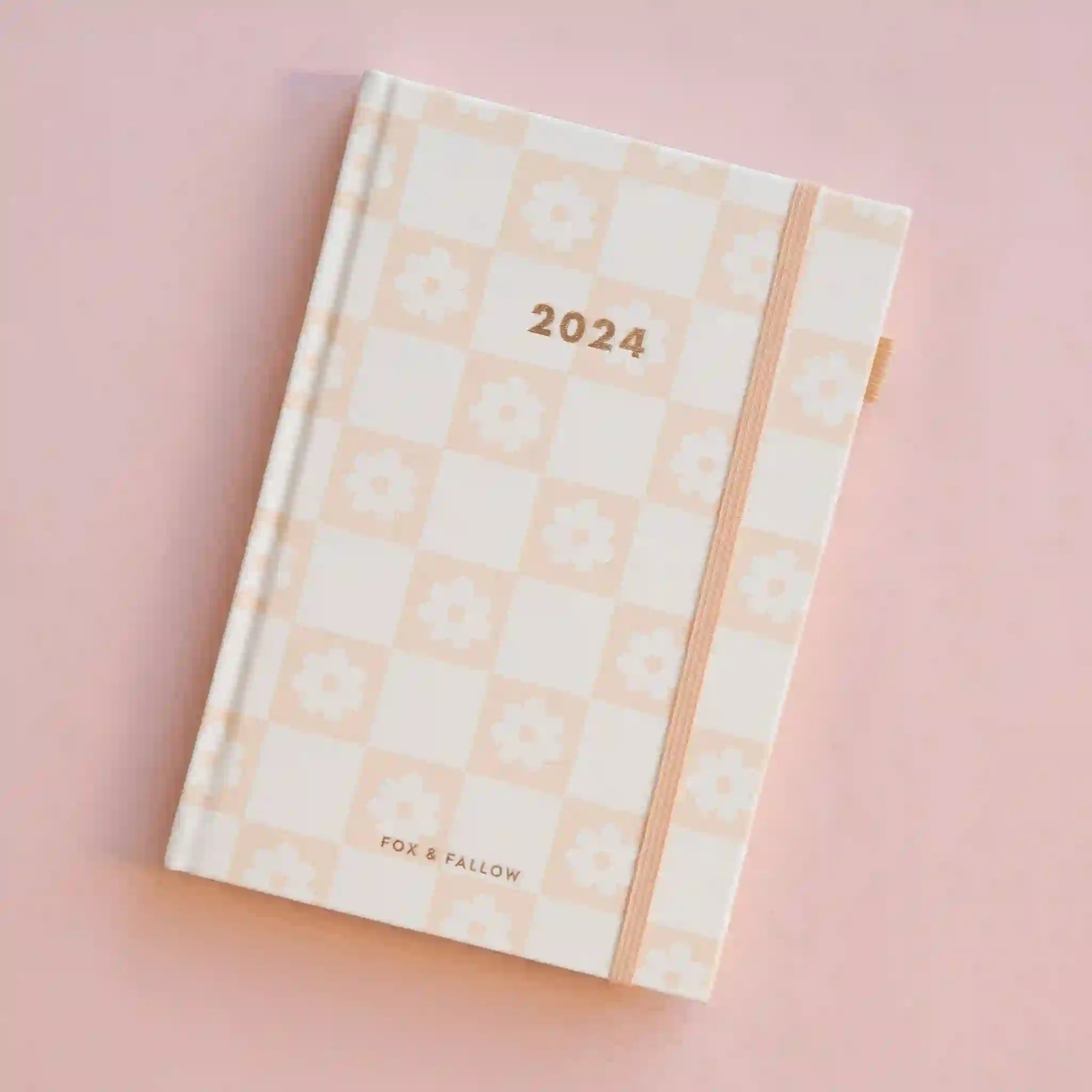 On a pink background is a light pink and ivory checkered planner with gold foiled &quot;2024&quot; text in the center along with a light pink elastic loop for keeping shut.