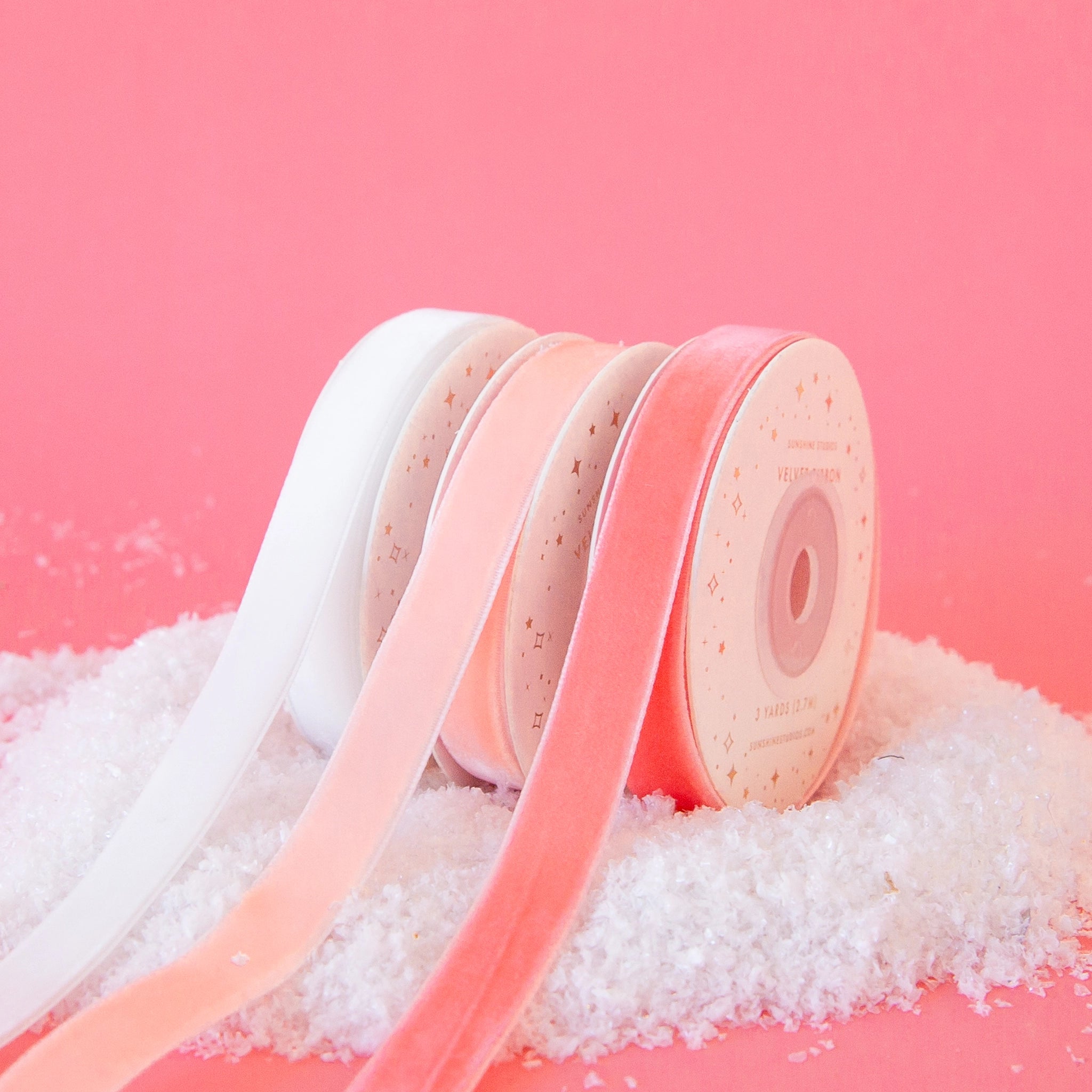 On a pink background is three spools of velvet ribbon in three shades. From left to right the colors are white, light pink and peach.