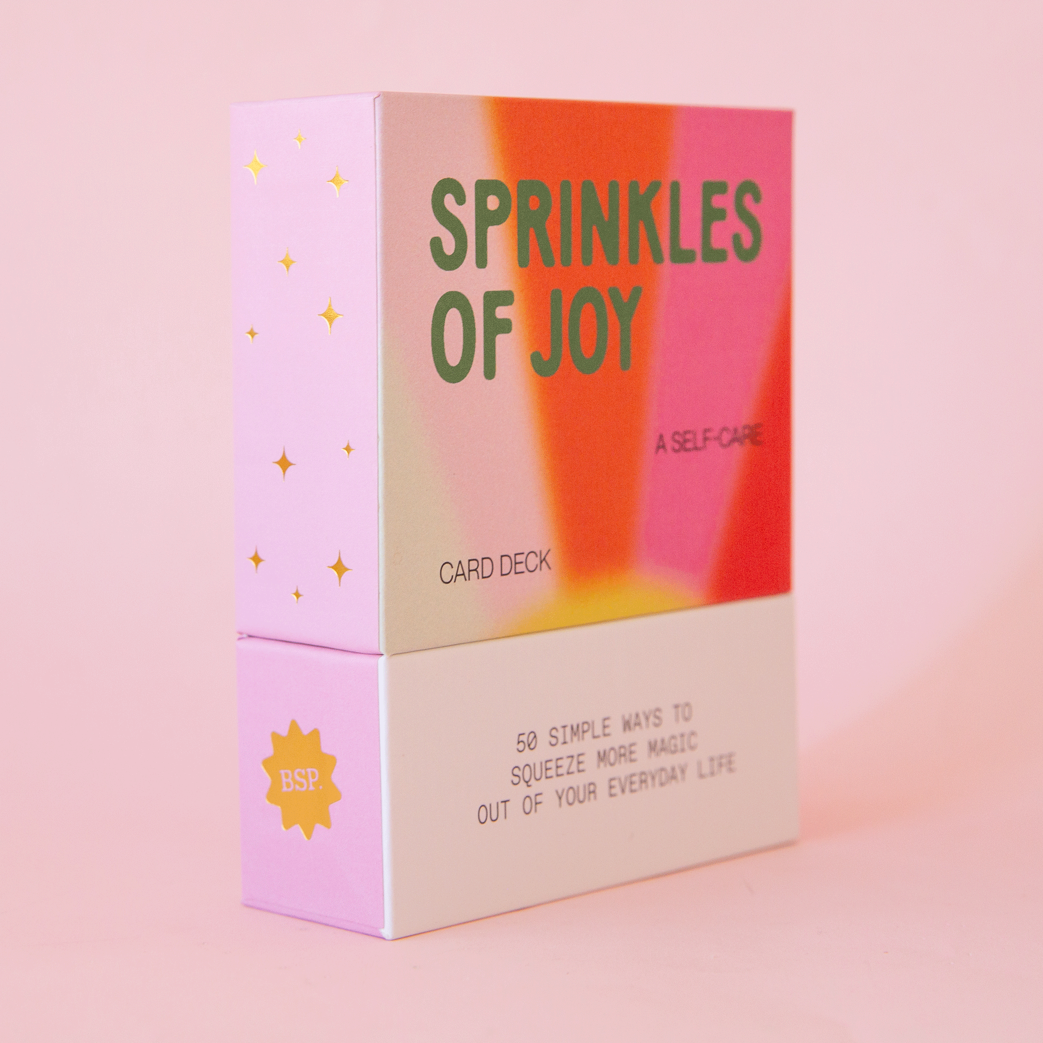 On a pink background is a deck of cards with a multicolored cover and text that reads, &quot;Sprinkles Of Joy, A Self-Care Card Deck&quot;.