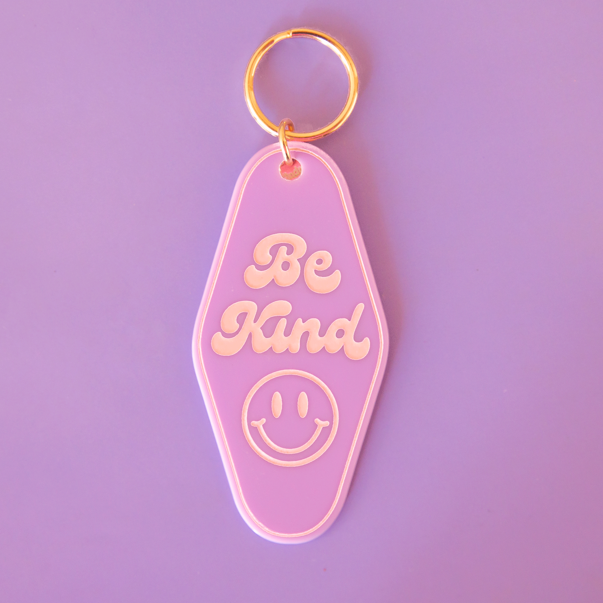On a purple background is a diamond shaped keychain in a lilac shade with gold detailing, a smiley face and text that reads, "Be Kind".