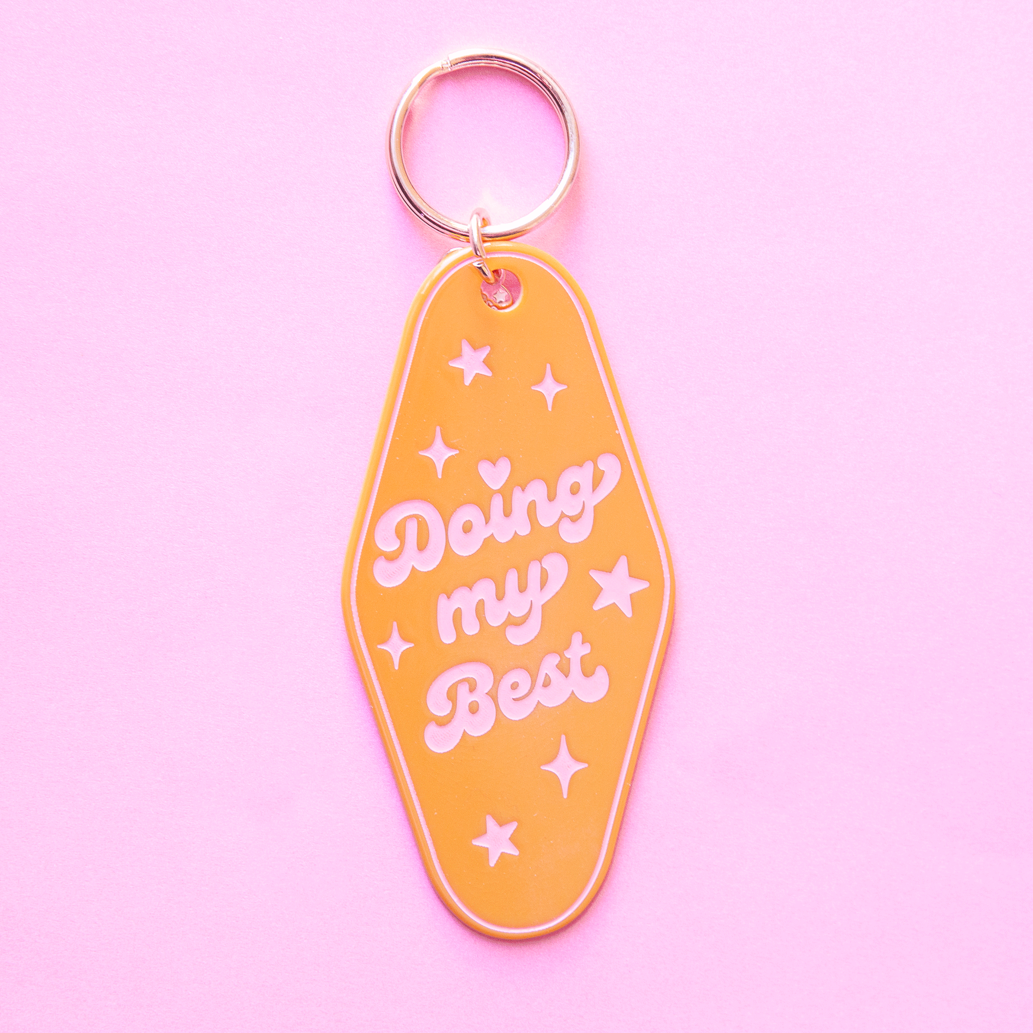 On a purple background is a mustard yellow diamond shaped keychain with a gold loop and pink text that reads, &quot;Doing my Best&quot; surrounded by star shapes and sparkles.