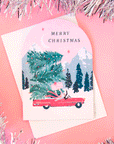 On a pink background is a pink arched card with a snowy mountain background with a red convertible stacked with Christmas trees.