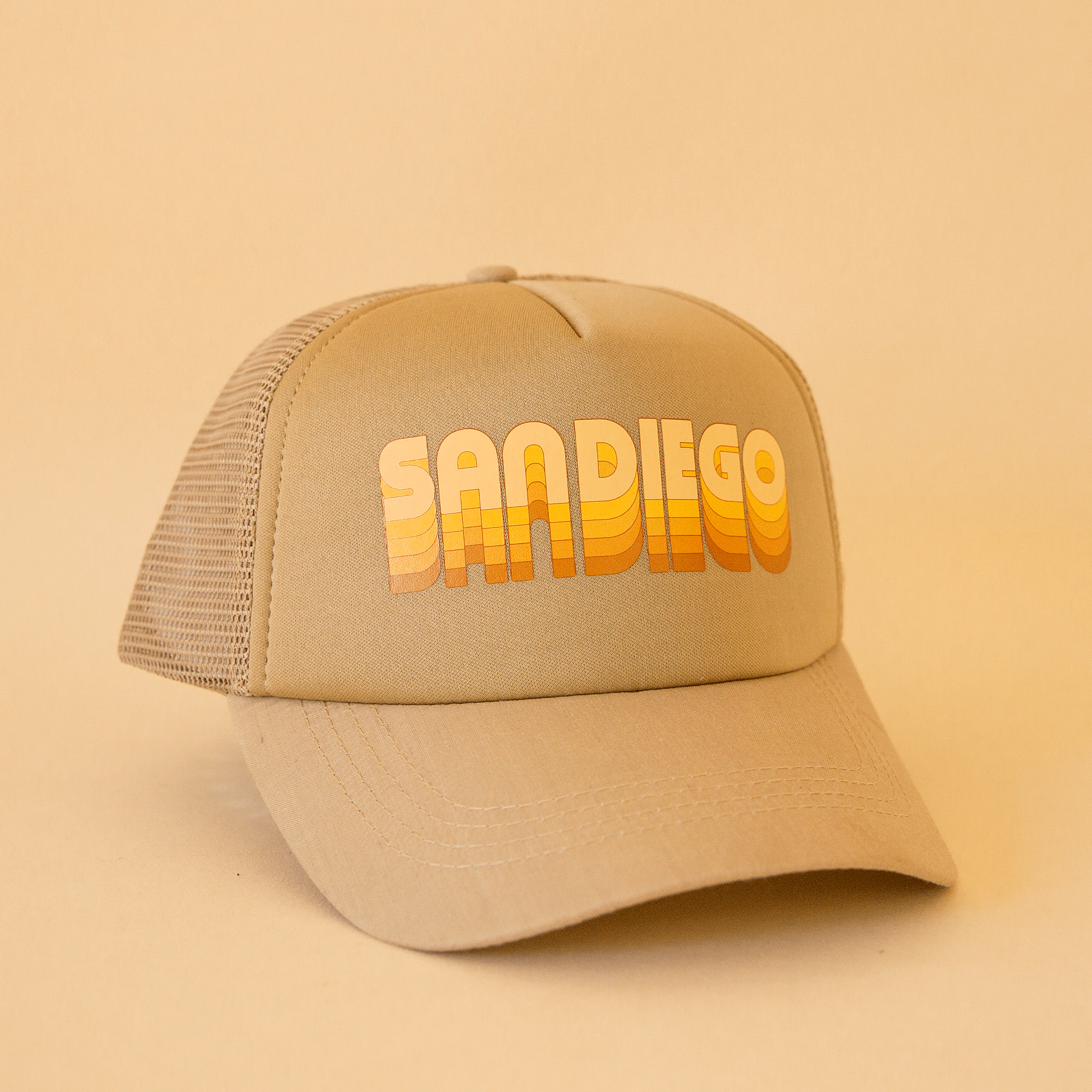 On a tan background is a tan trucker hat with yellow and orange gradient text that reads, &quot;San Diego&quot;.