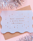 On a pink background is a wavy edged light blue card with gold foiled text that reads, "have yourself a merry little Christmas".