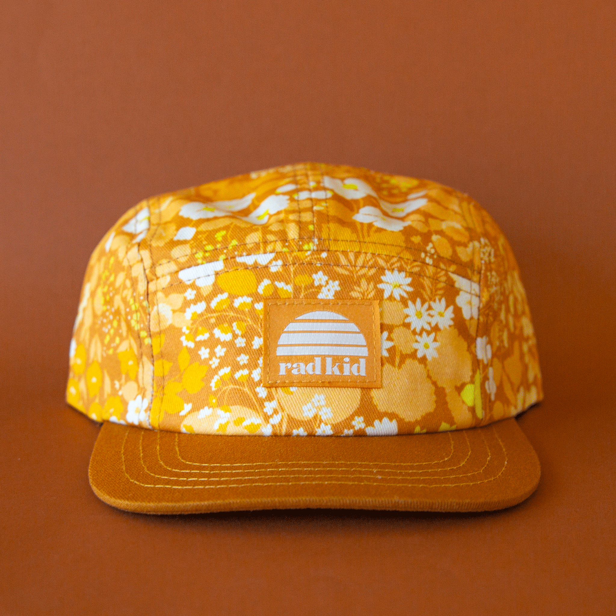On a burnt orange background is a yellow and brown floral print hat with a square label on the front that reads, "rad kid".