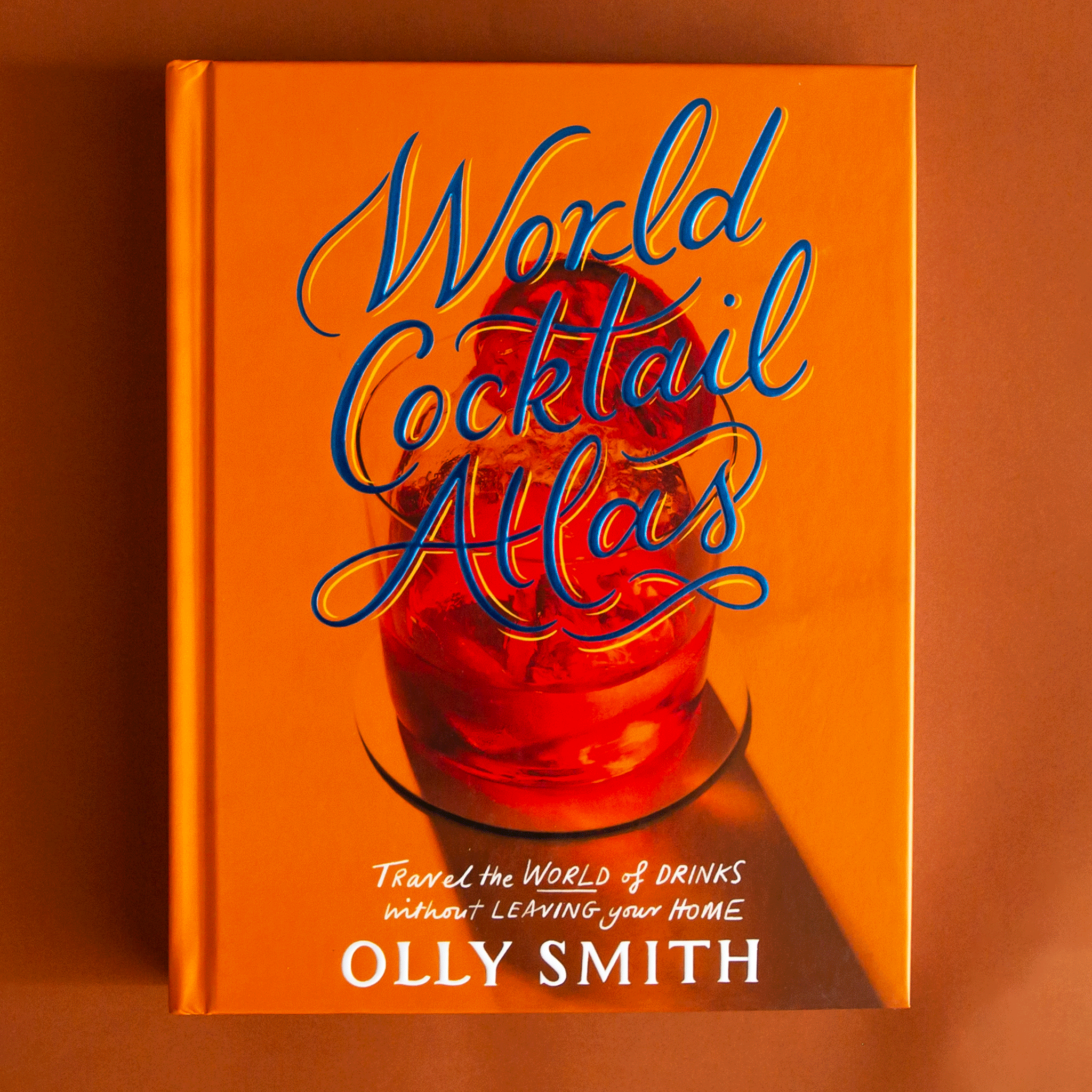 On an orange background is an orange cocktail book with a cocktail glass graphic along with blue text that reads, &quot;World Cocktail Atlas&quot;.