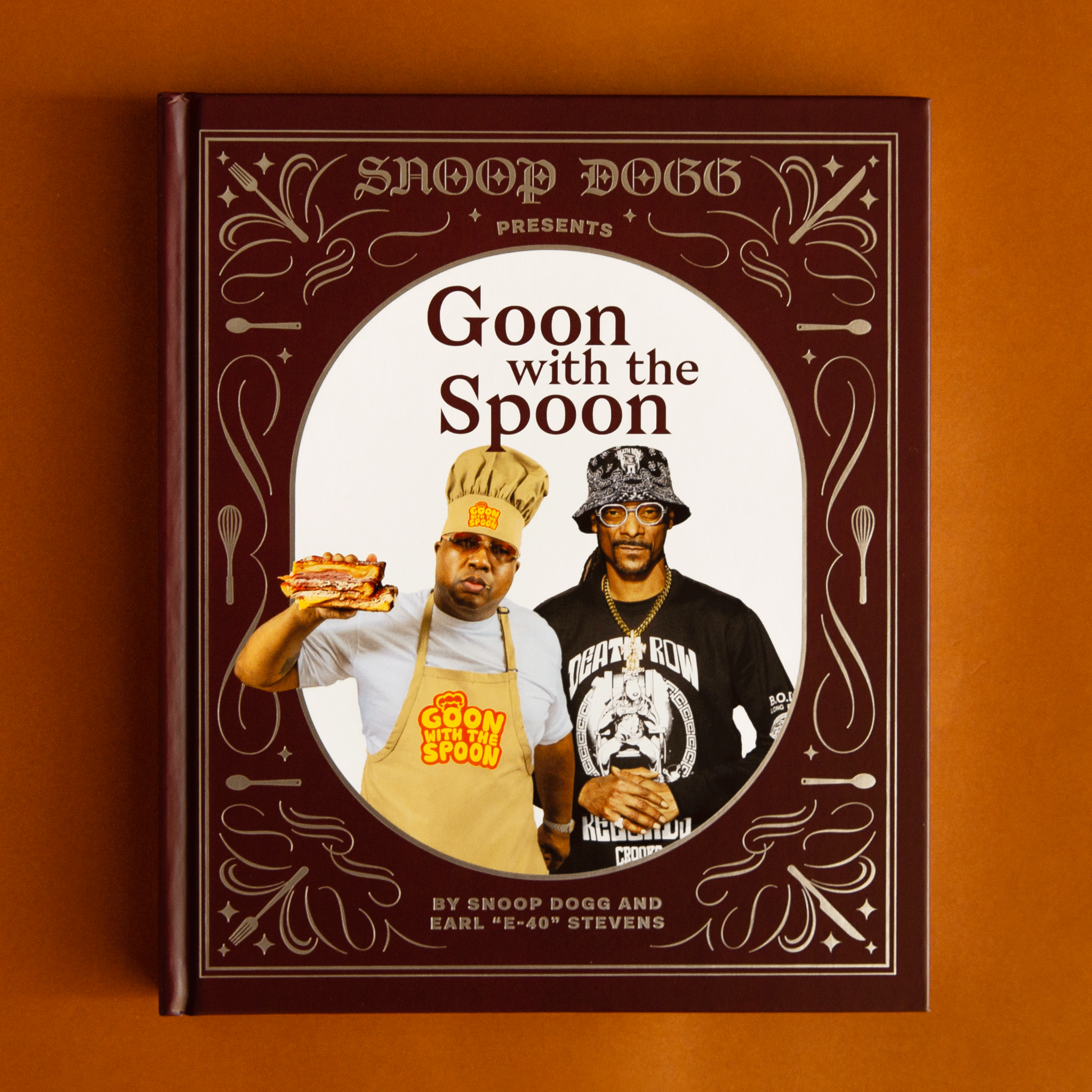 On a burnt orange background is a brown and gold book cover with a photo of Snoop Dog and E-40 with the title above it that reads, &quot;Goon with the Spoon&quot;.