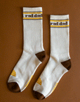 On a brown background is a pair of natural colored socks with brown heel and text that reads, "rad dad" along with brown, black and yellow accents.