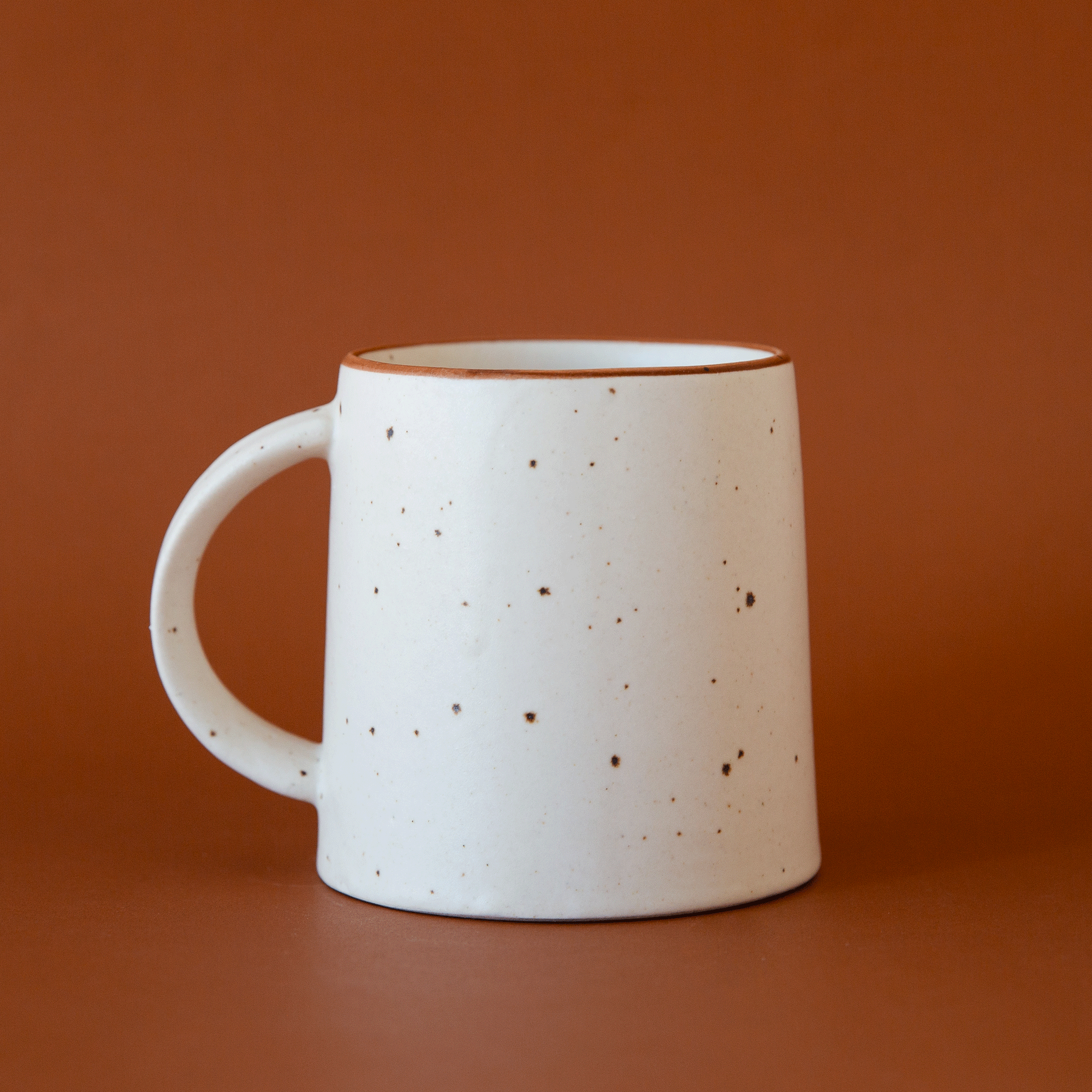 On a burnt orange background is a white and brown speckled ceramic mug with a curved handle.