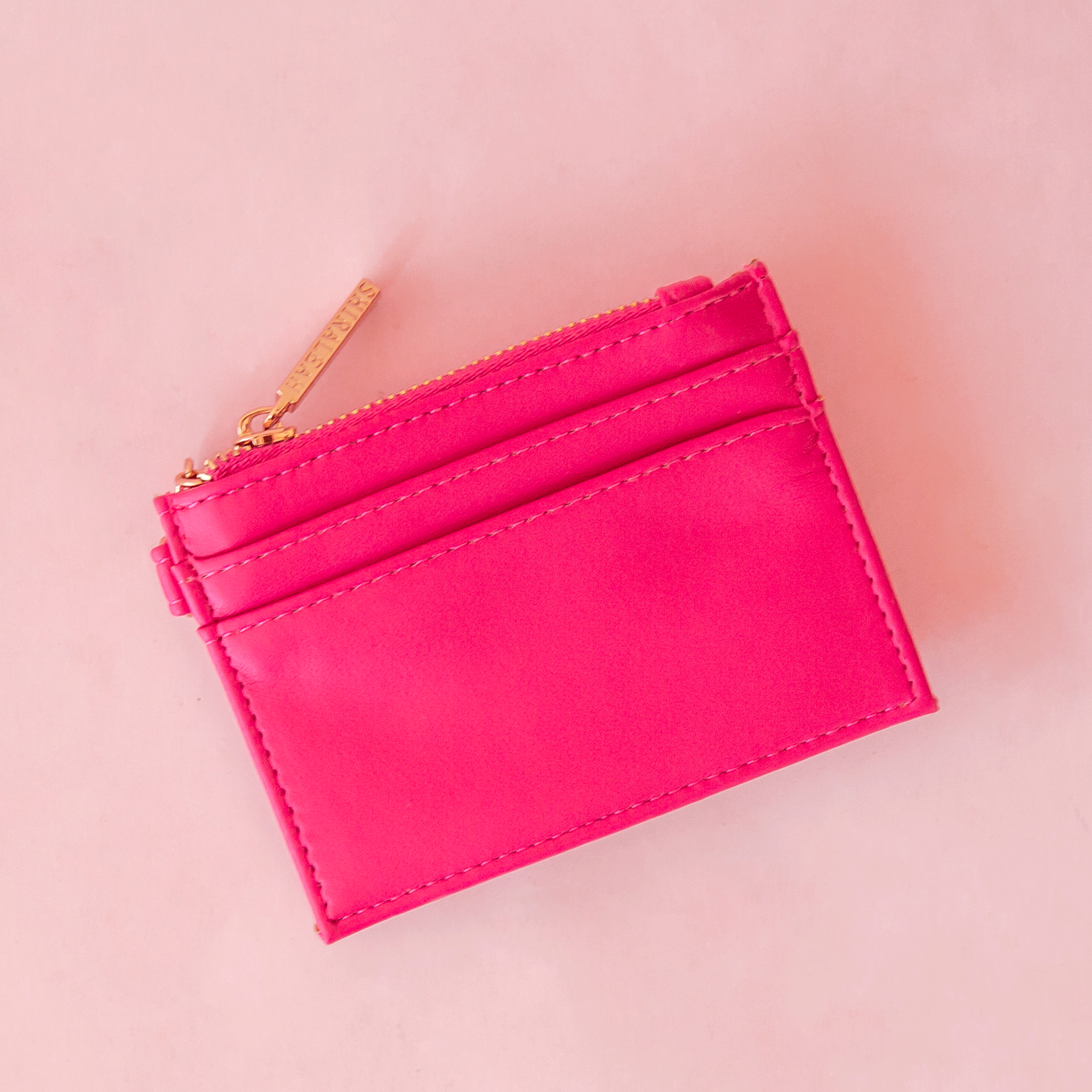 On a pink background is a hot pink card case with a gold zipper and details.
