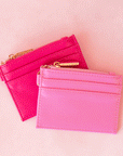 On a pink background is a bubble gum pink card case next to a hot pink version with a gold zipper and details.