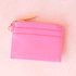 On a pink background is a bubble gum pink card case with a gold zipper and details. 