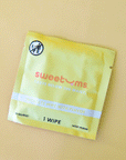 On a yellow background is a yellow packet of a single use feminine wipe with text on the front that reads, "sweetums taste below the waist".