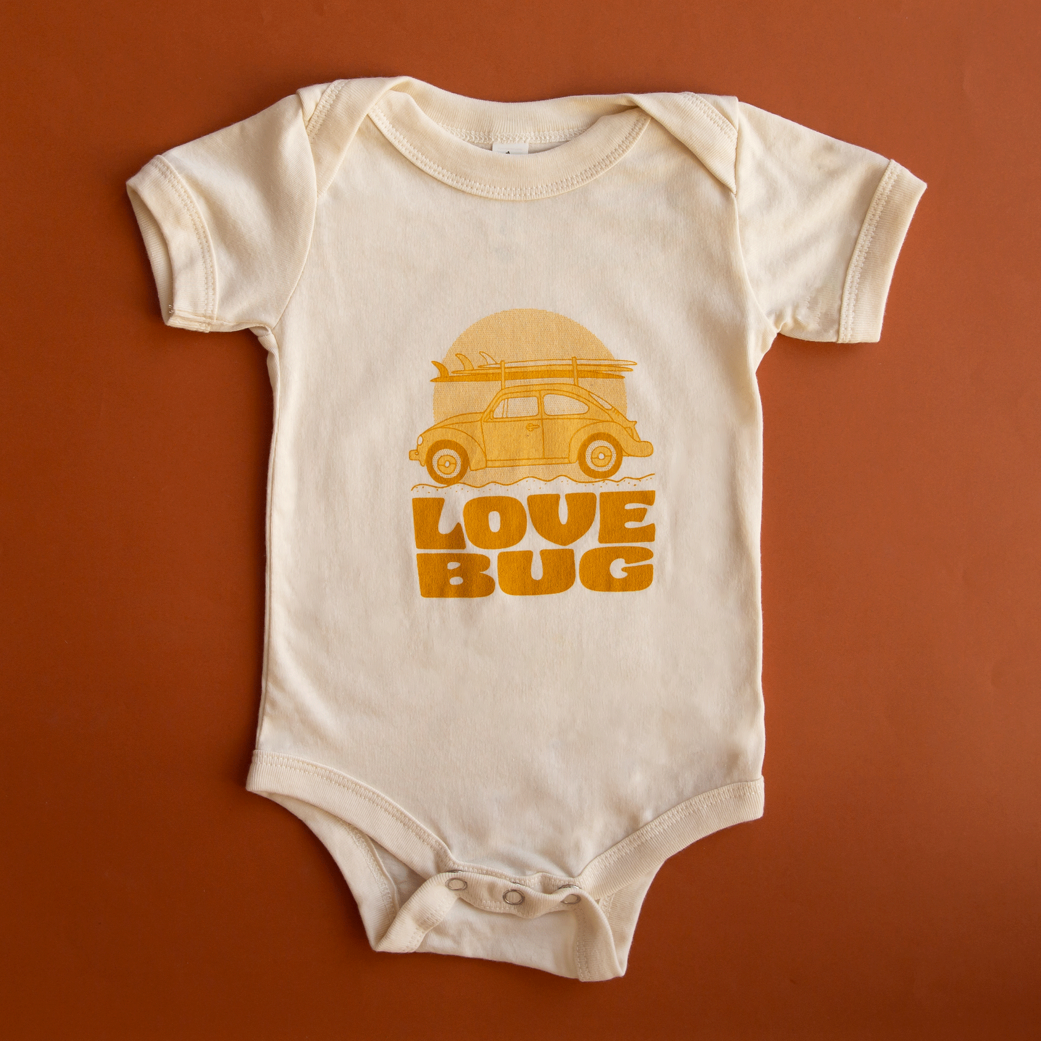 On a burnt orange background is an ivory short sleeve children&#39;s onesie with a yellow VW bug with a surfboard graphic with orange letters underneath that reads, &quot;Love Bug&quot;. 