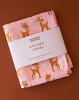 On a brown background is a pink and waffle knit kitchen towel with a brown retro reindeer pattern.