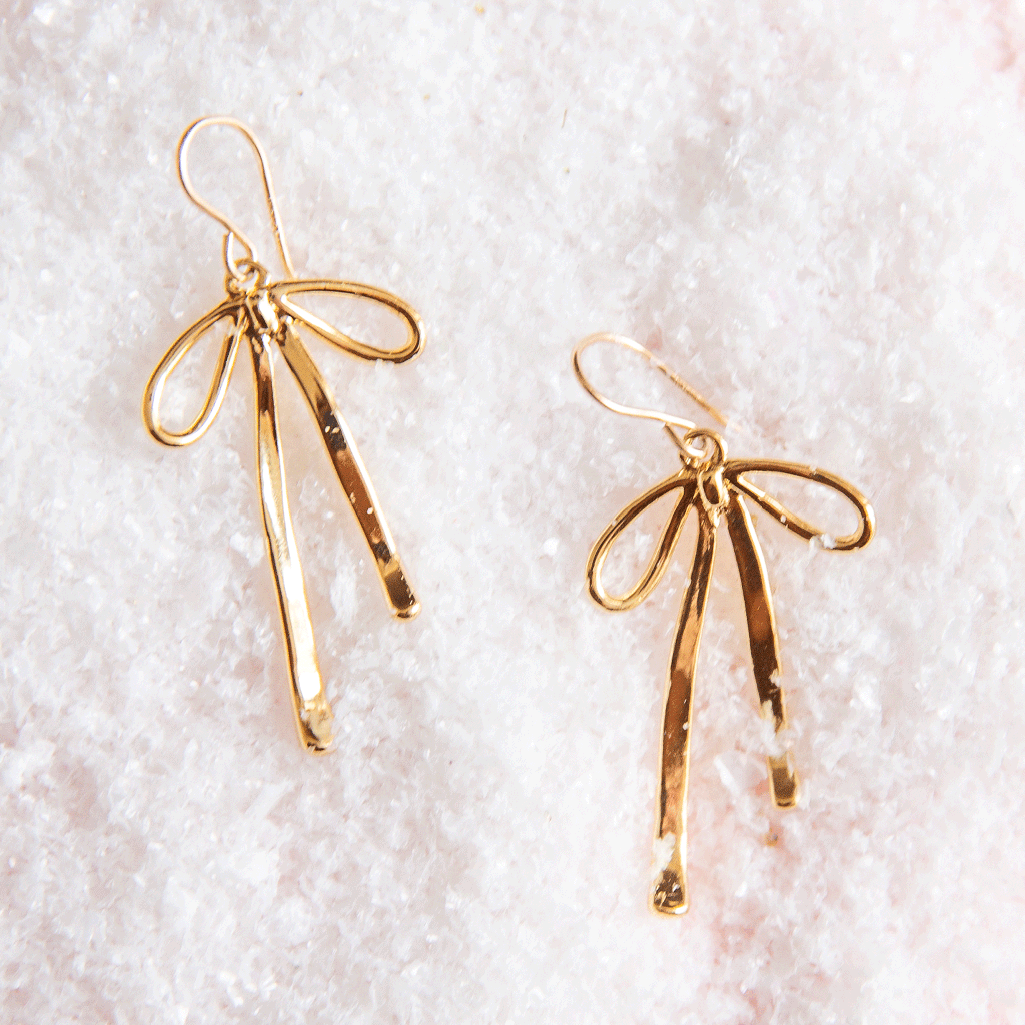 On a pink snowy background is a pair of gold bow shaped earrings.