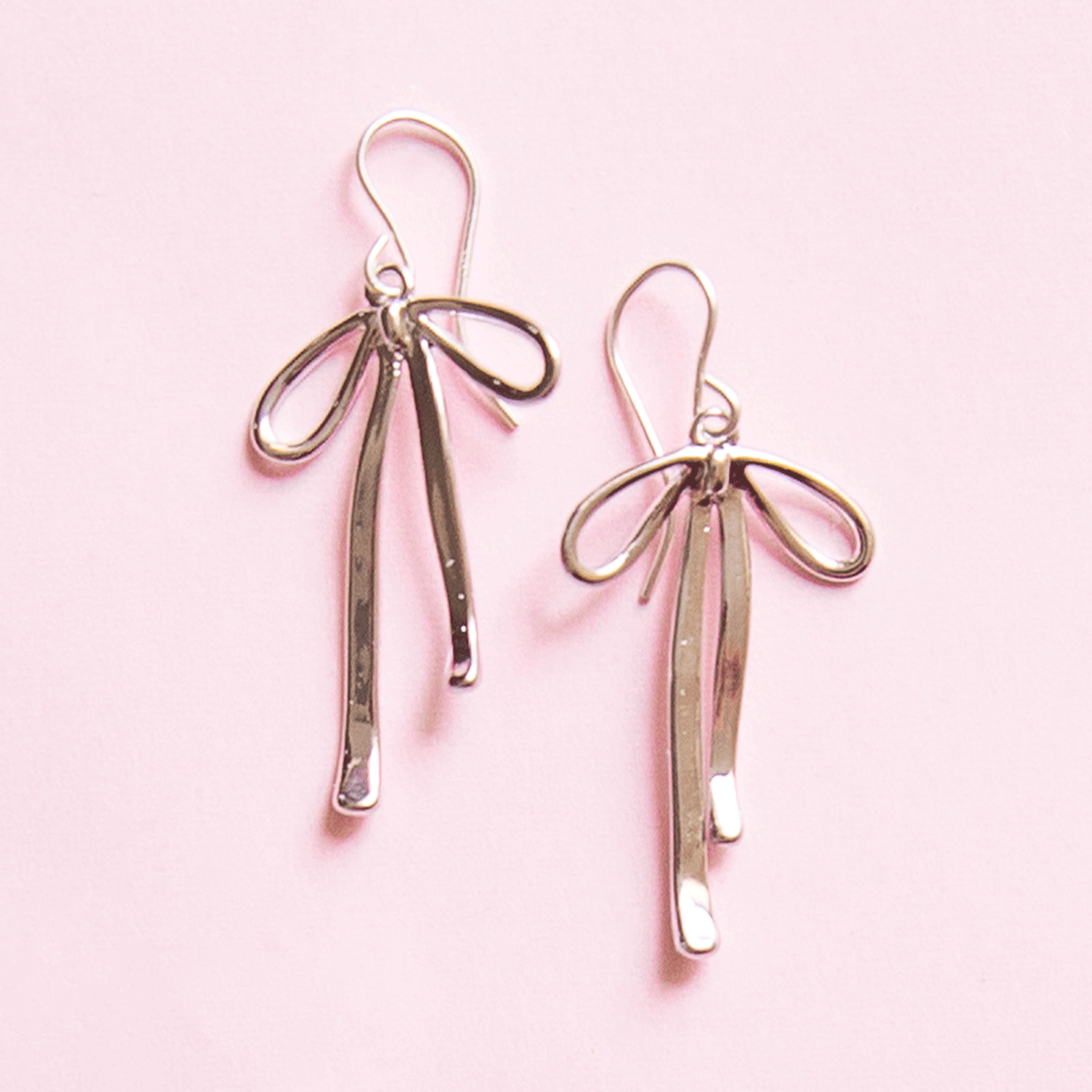 On a pink background is a pair of silver bow shaped earrings.