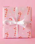 A single sheet of gift wrap with a blush background and a repeating red and white candy cane print. The gift wrap is reversible and the other side features a light purple / lavender background with an all over red and white candy cane design but the candy canes are slightly smaller than the reverse. Present is wrapped with a silver tinsel ribbon and surrounded by snow.