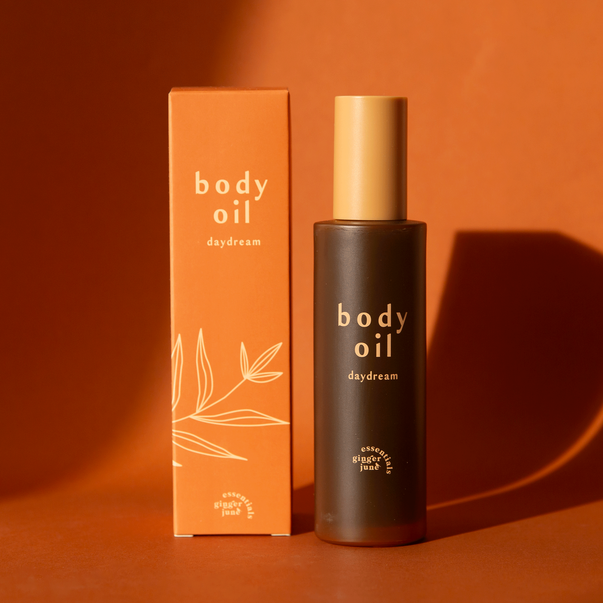 On an orange background is a bottle of body oil with a tan cap and an orange box packaging along with text on the front that reads, &quot;body oil daydream&quot;.