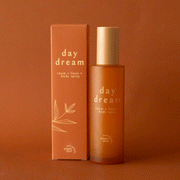 On a burnt orange background is an orange packaged spray with text that reads, "day dream room linen body spray".