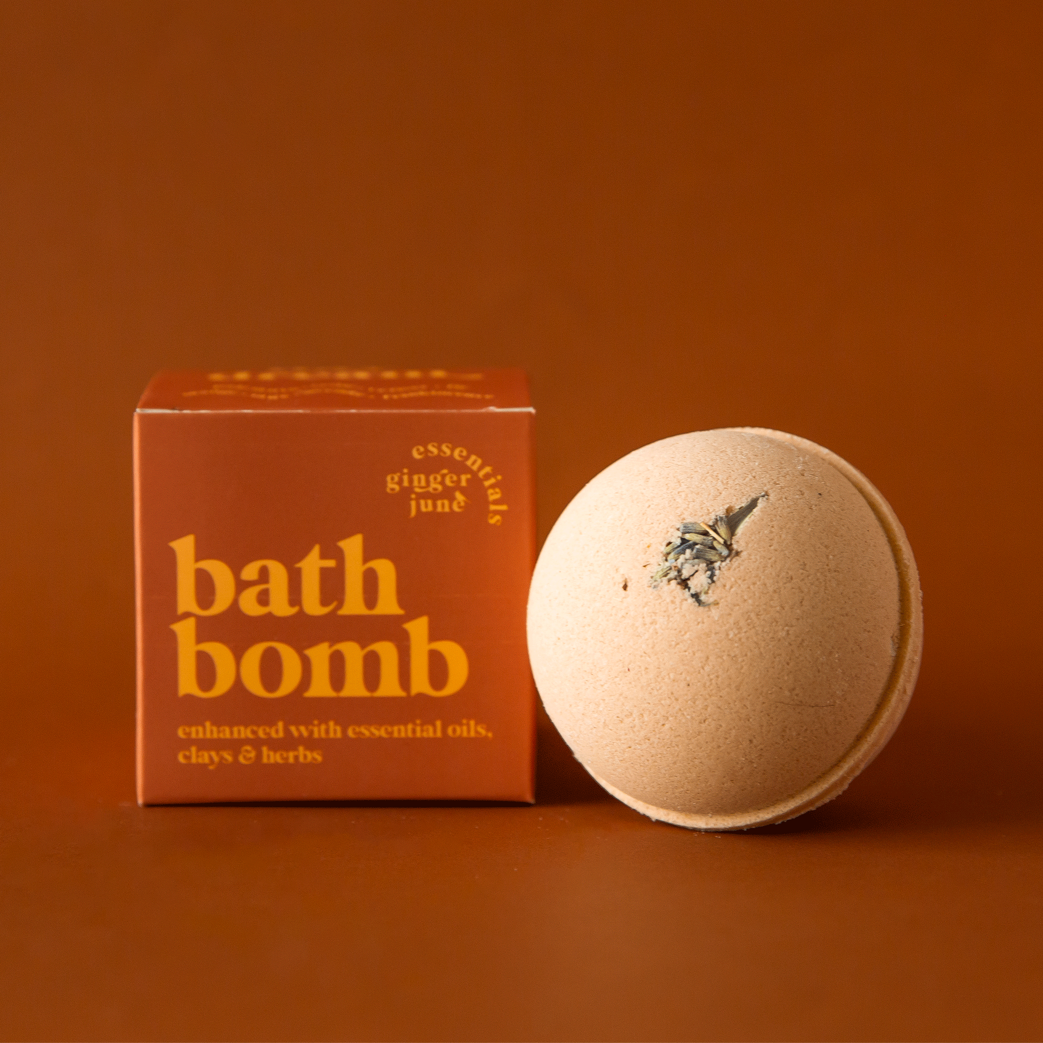 On a burnt orange background is a round bath bomb with an orange hue and a orange box with yellow text that reads, &quot;bath bomb enhanced with essential oils, clays &amp; herbs&quot;.