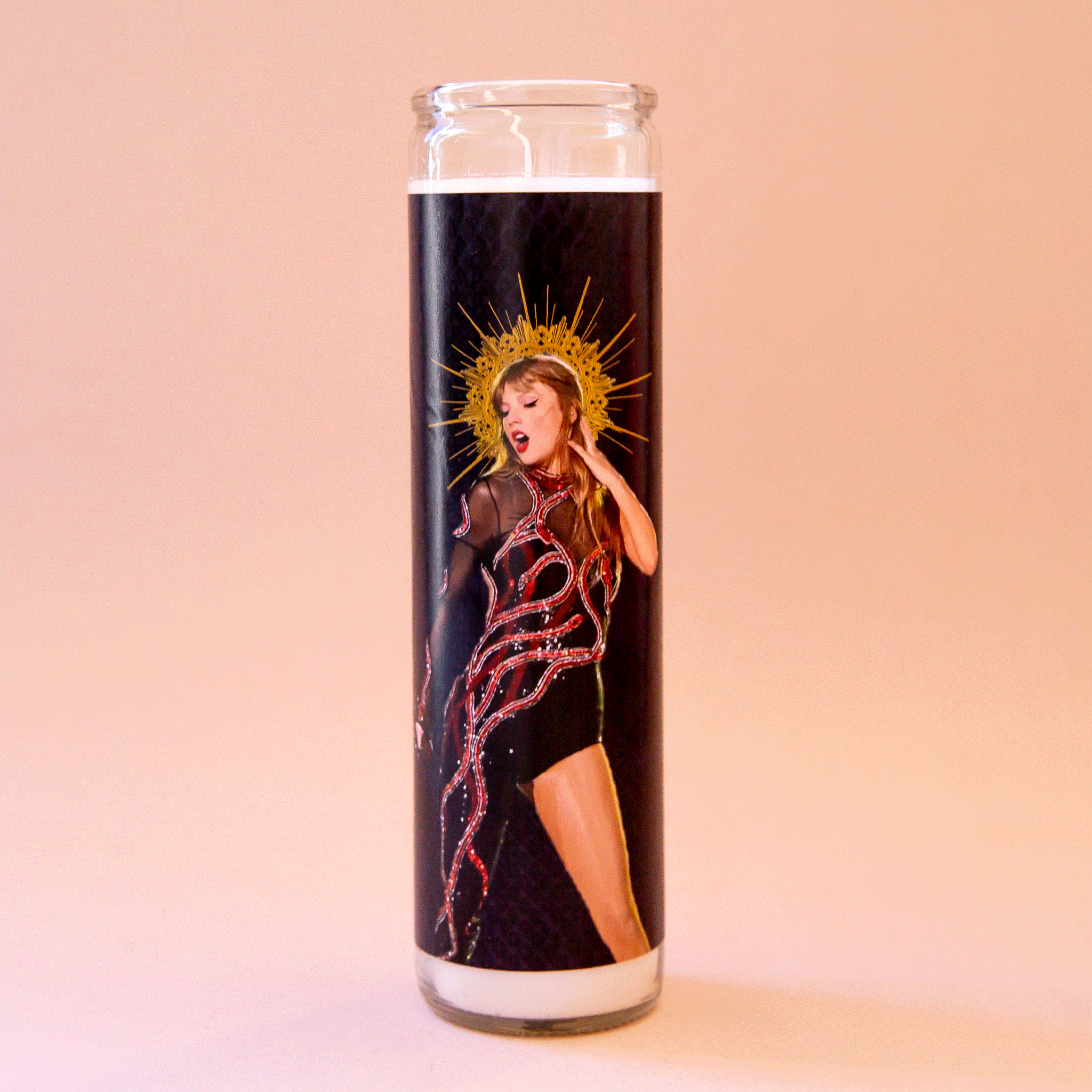 On a peach background is a thin prayer candle with a black label featuring a picture of Taylor swift in her reputation outfit on a black background. 