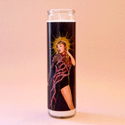 On a peach background is a thin prayer candle with a black label featuring a picture of Taylor swift in her reputation outfit on a black background. 