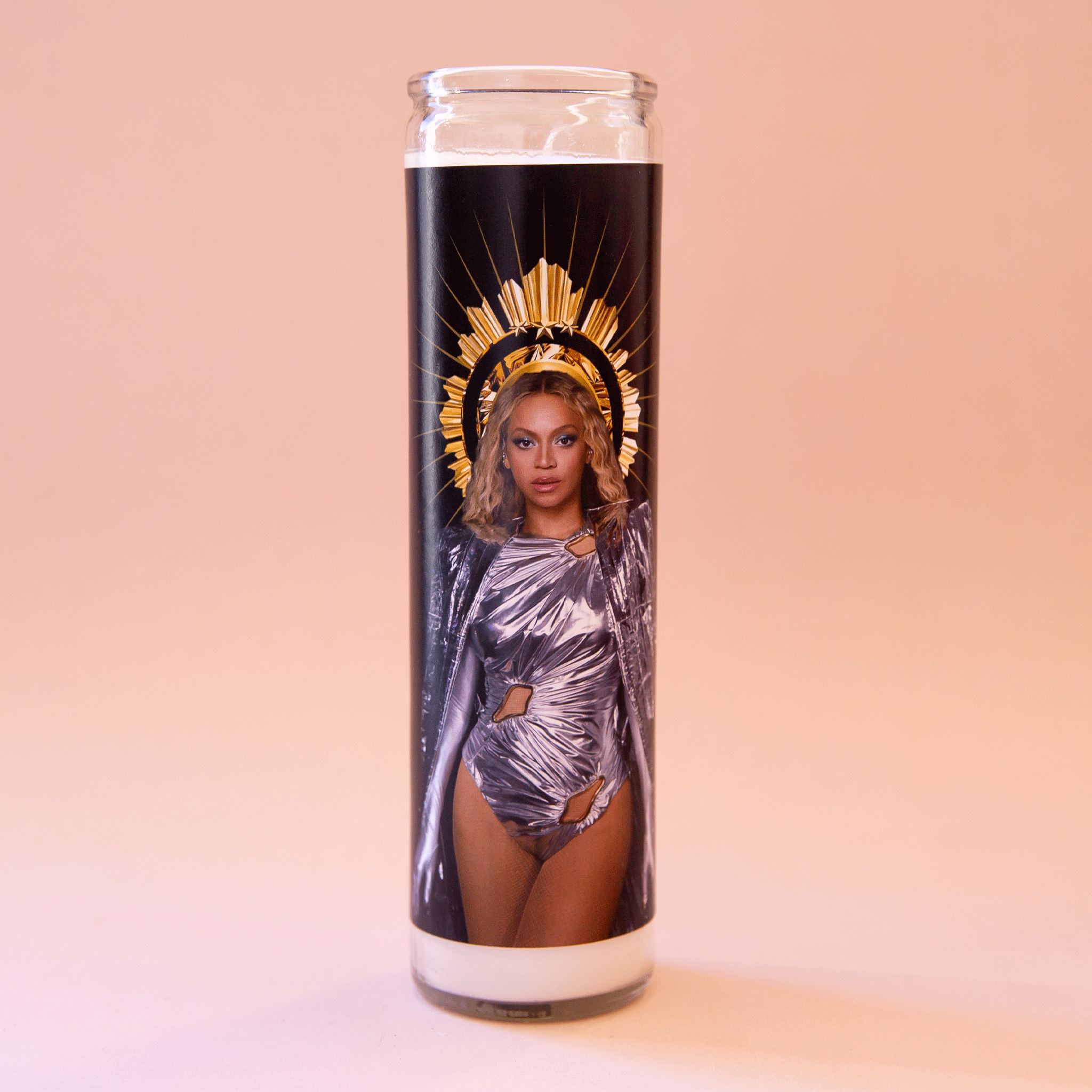 On a peach background is a prayer candle with a photo of Beyonce in a silver outfit with a crown and in front of a black background. 