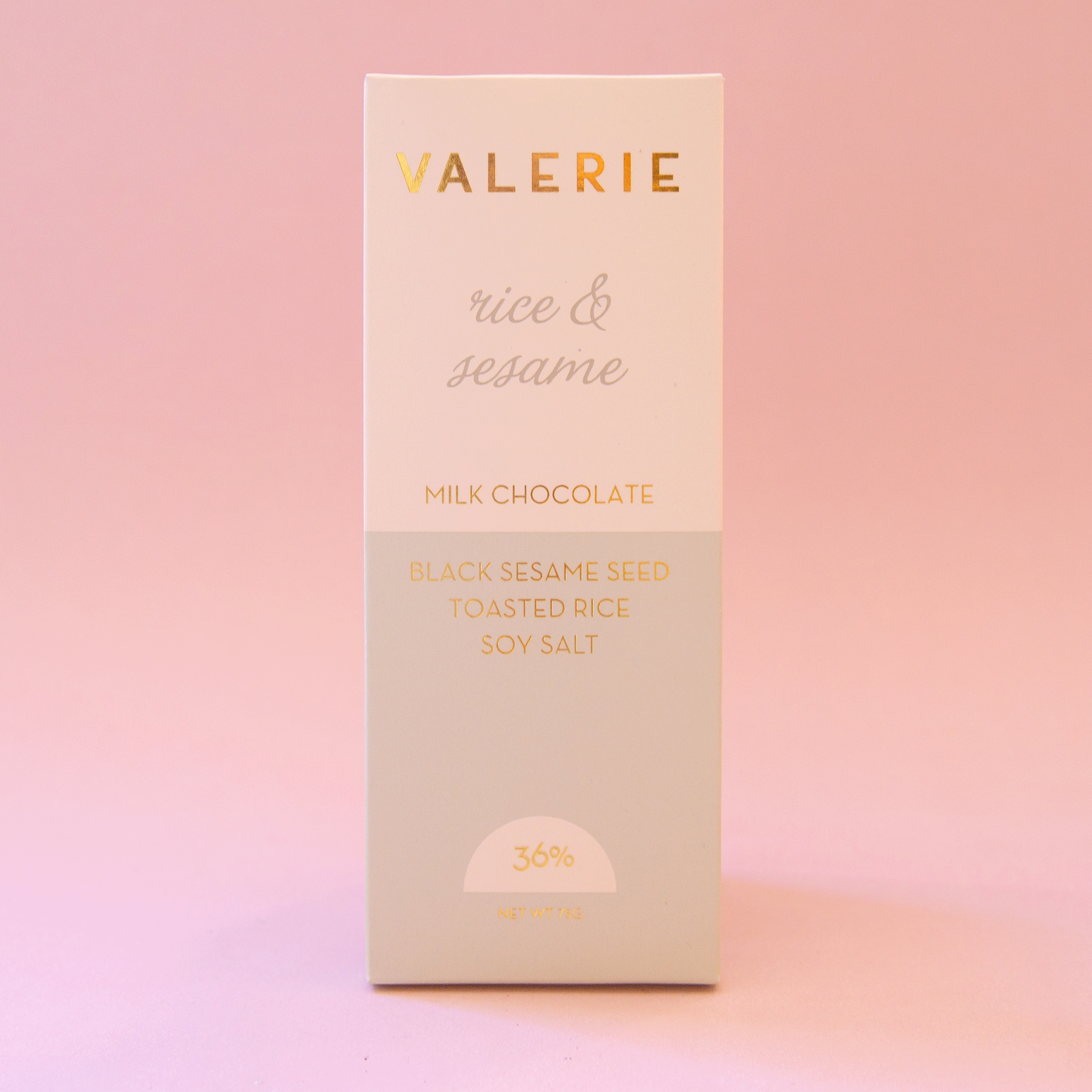 On a pink background is a neutral colored bar of chocolate with gold foiled text that reads, &quot;Valerie rice &amp; sesame Milk Chocolate&quot;.